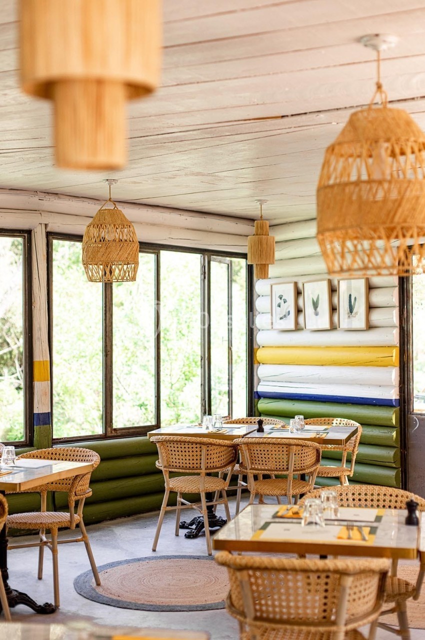 Salle de restaurant lumineuse avec mobilier en rotin, grandes fenêtres et décorations en bois naturel.