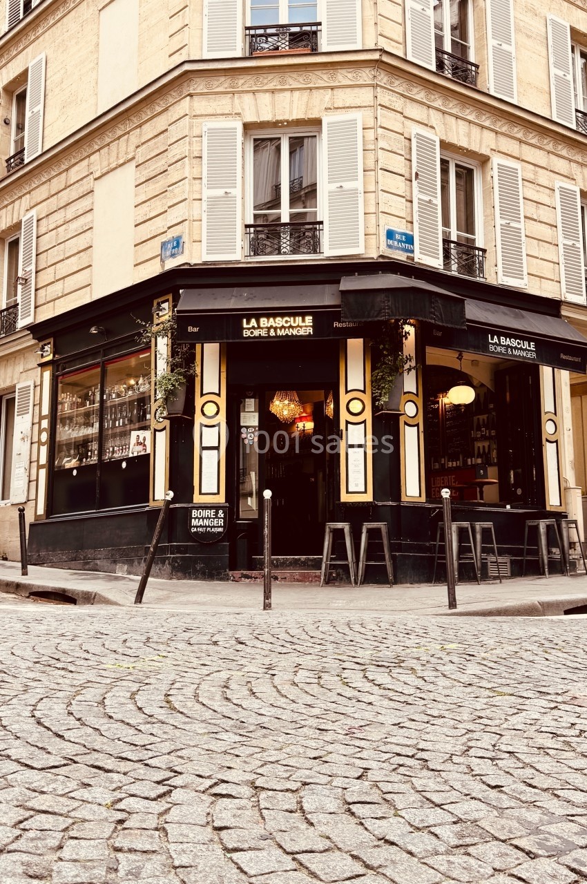 Façade d'un café-restaurant d'angle avec terrasse et grandes vitrines, situé sur une rue pavée.