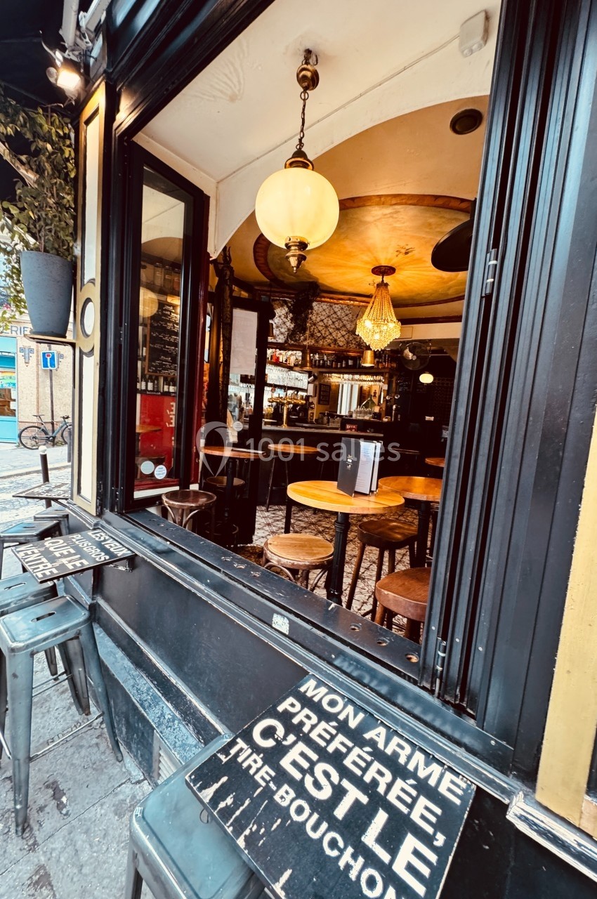 Vue d'un café avec une fenêtre ouverte, des tables en bois à l'intérieur et un panneau humoristique au premier plan.