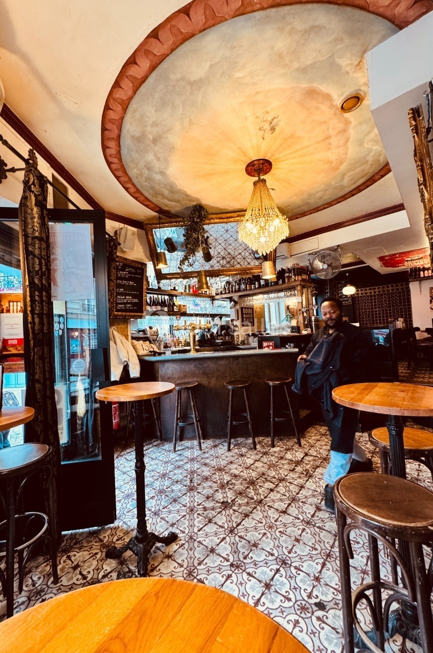Intérieur d'un café avec un bar en bois, des tables rondes, un plafond décoré et un lustre central.