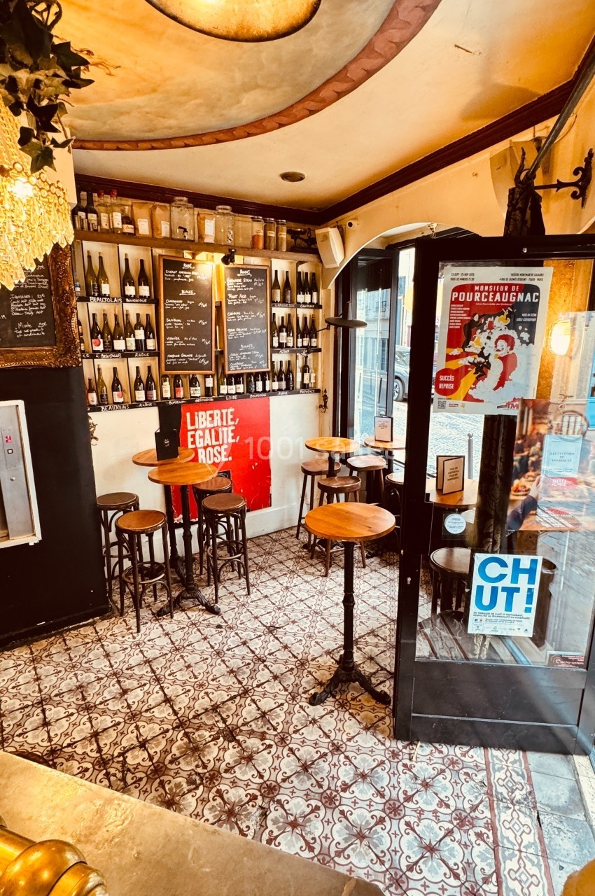 Intérieur d'un petit bar avec des tables hautes, des étagères remplies de bouteilles et des affiches sur les murs.