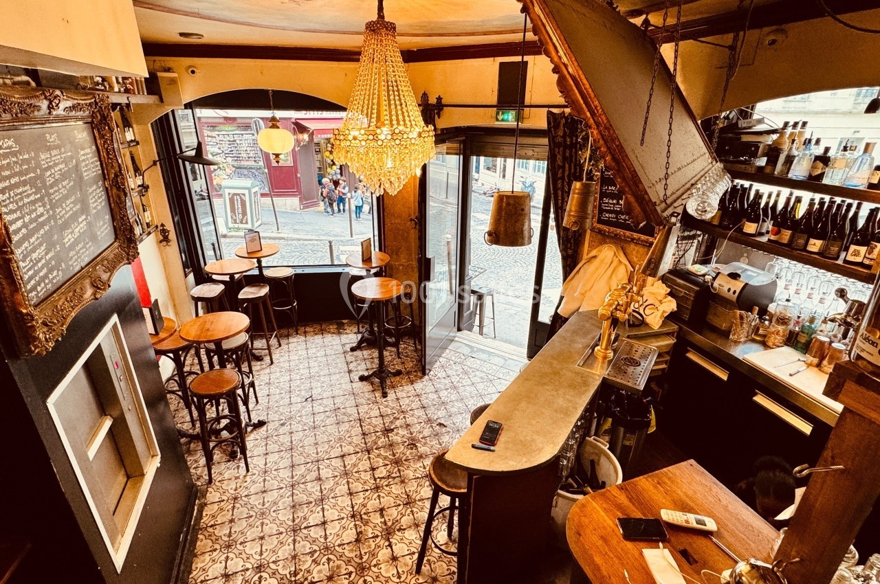 Intérieur d'un café avec comptoir en bois, tables hautes, lustre central et vue sur une rue pavée par la vitrine.