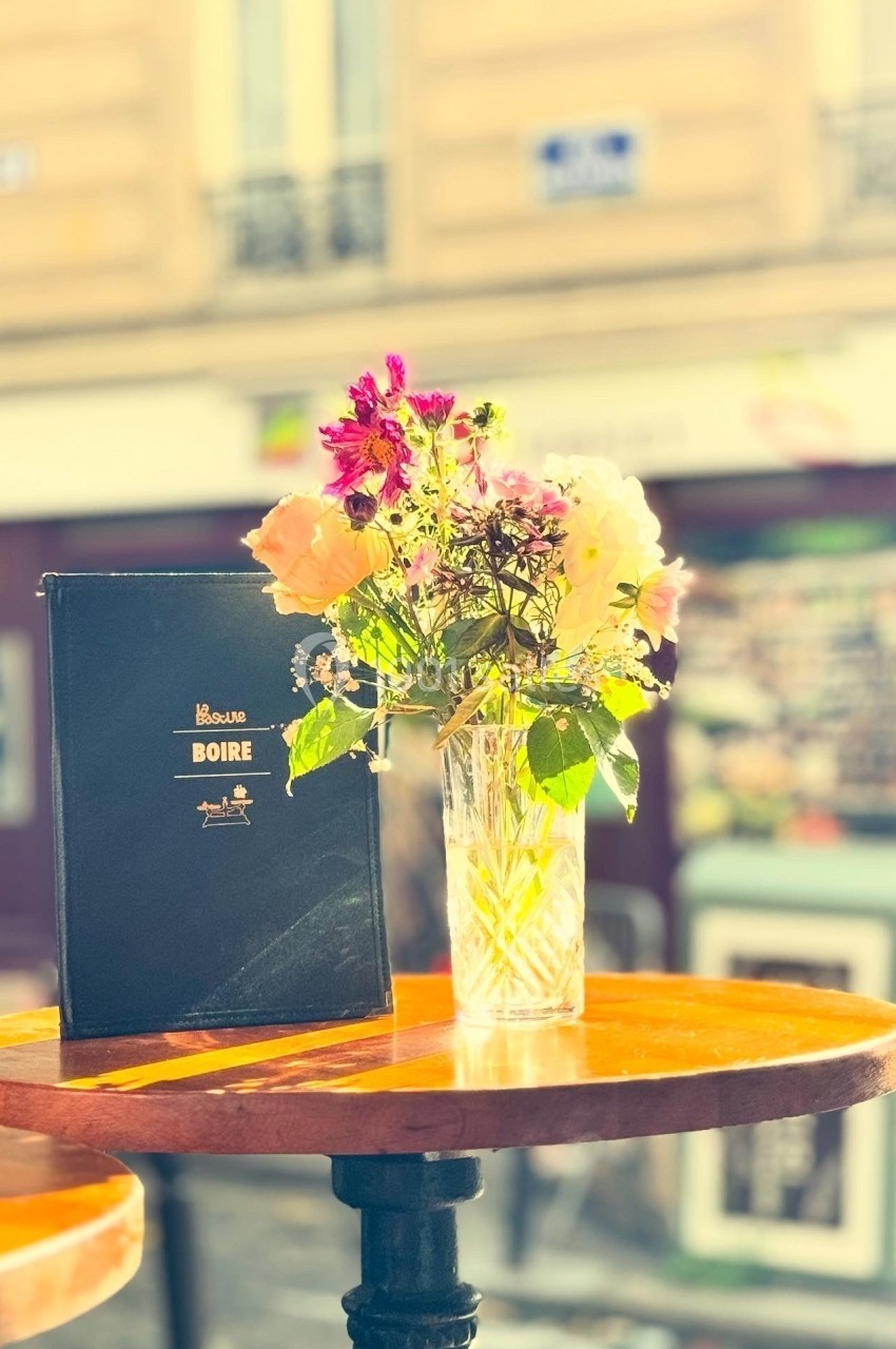 Un vase de fleurs colorées et un menu posé sur une table en bois, en extérieur devant une façade floue.