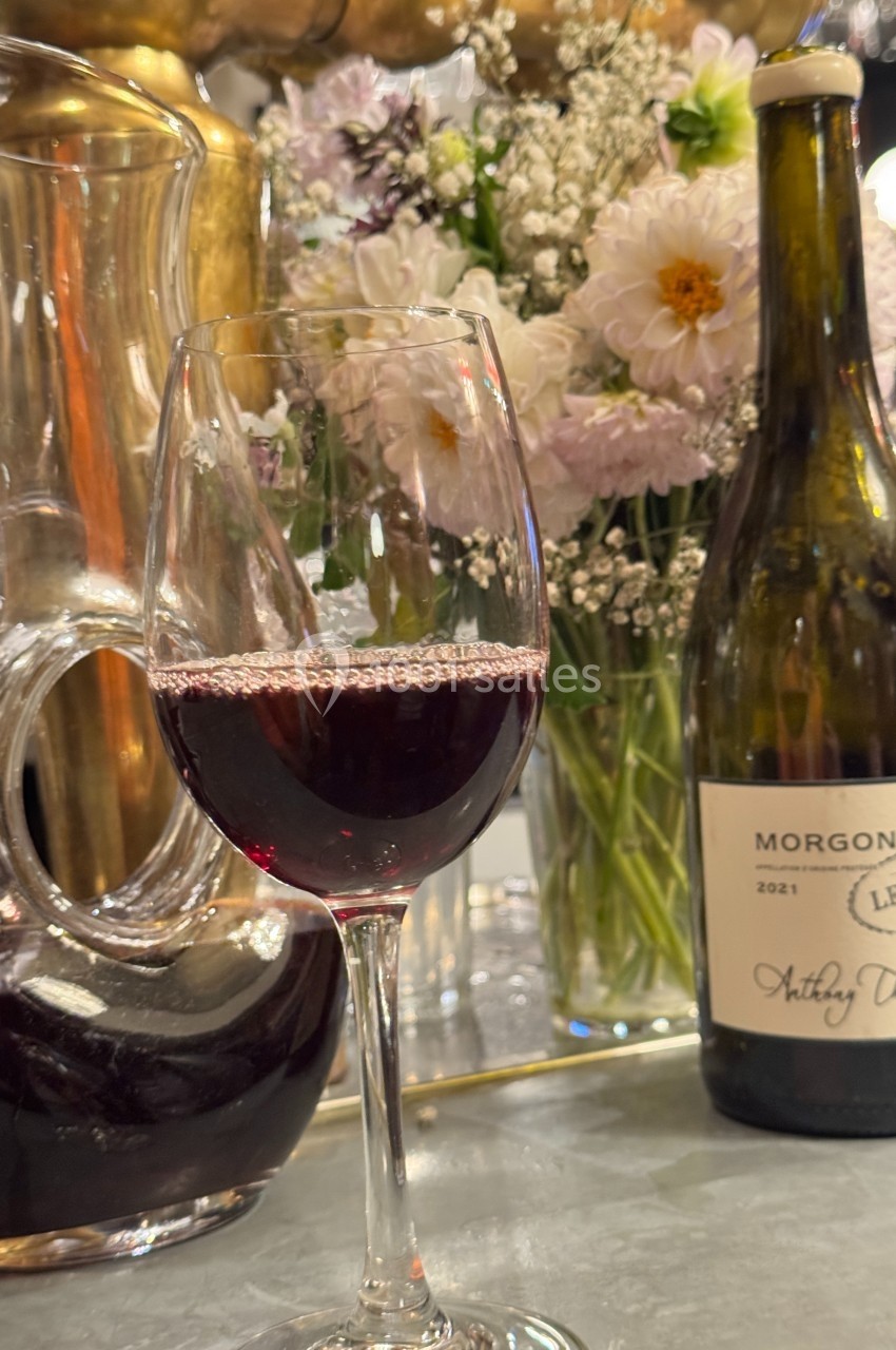 Verre de vin rouge devant une carafe, une bouteille de Morgon 2021 et un bouquet de fleurs en arrière-plan.