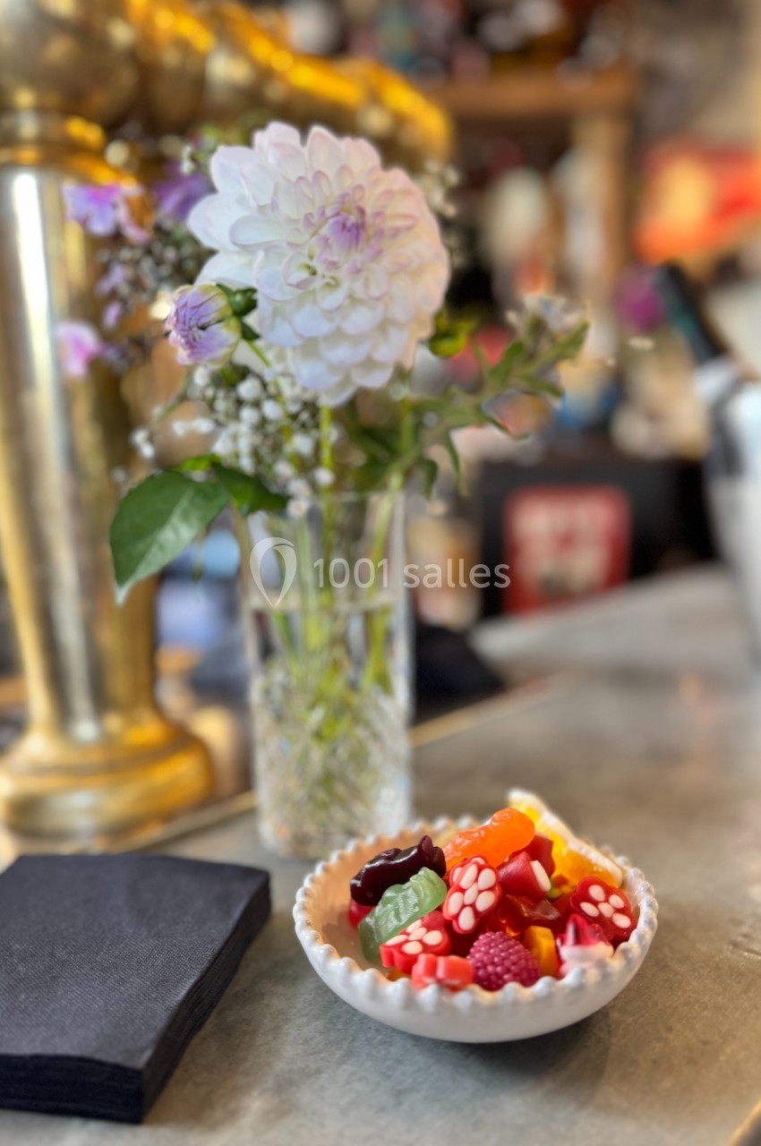 Bol de bonbons colorés posé sur un comptoir, avec un vase de fleurs blanches et violettes en arrière-plan.
