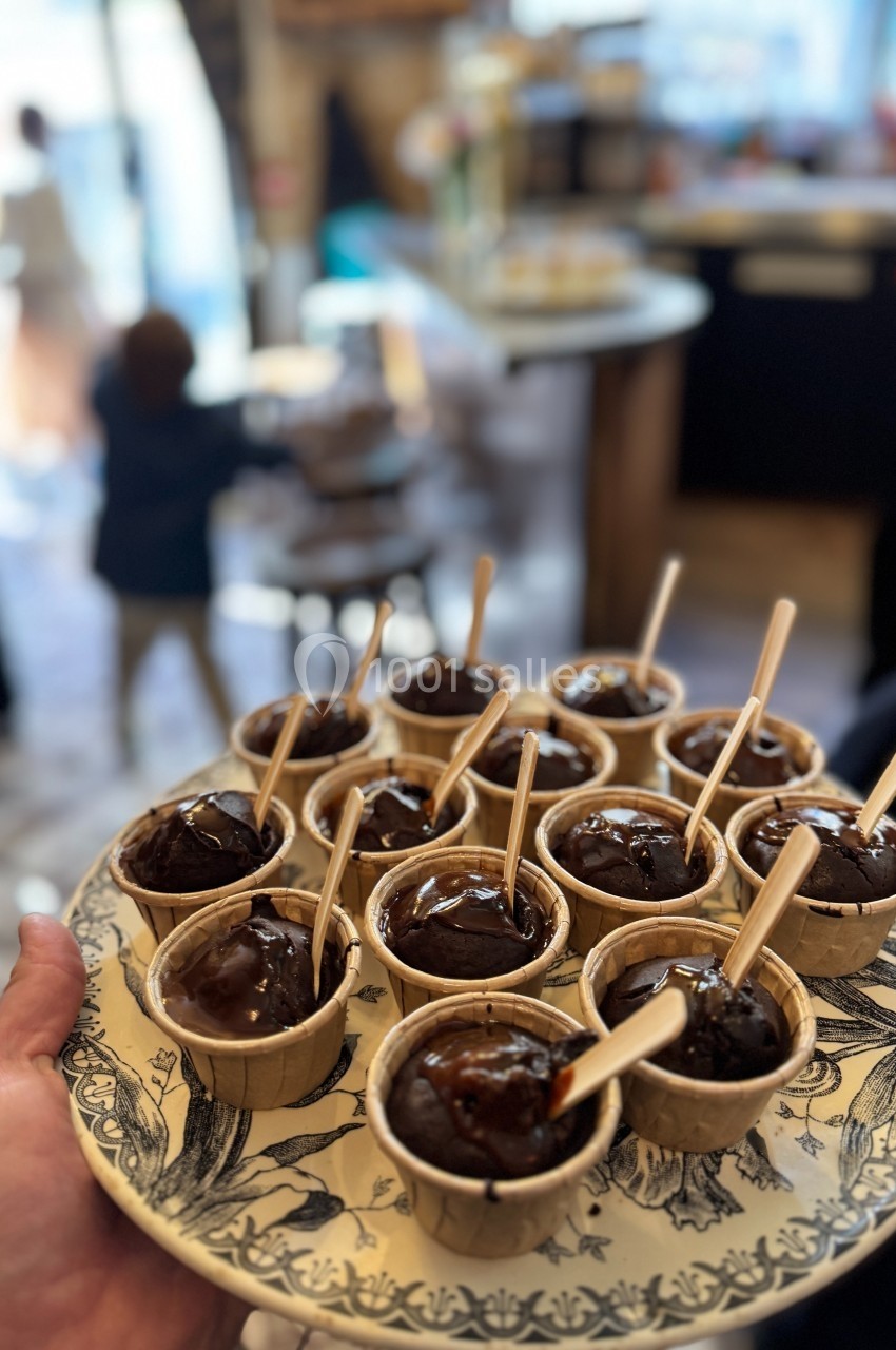 Plateau de petits muffins au chocolat servis dans des moules en papier, avec des cuillères en bois, dans un café animé.