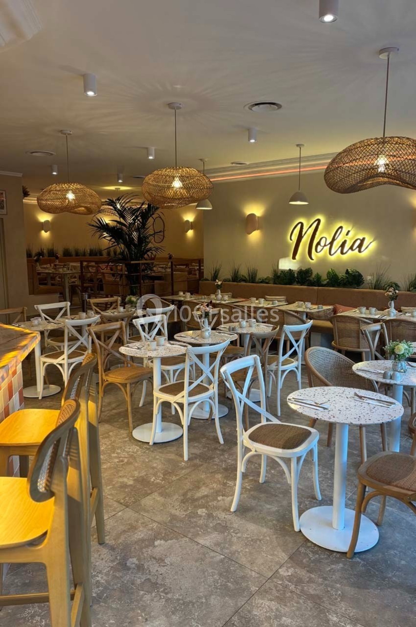 Salle de restaurant lumineuse avec mobilier en bois clair, tables blanches et enseigne lumineuse ’Nolia’ au mur.