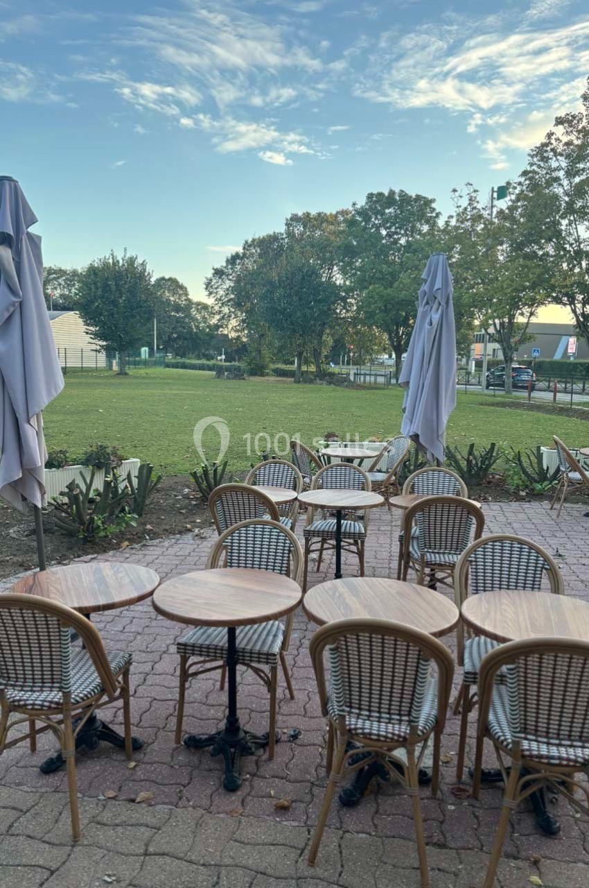 Terrasse extérieure avec tables et chaises en rotin, parasols fermés, donnant sur un espace vert et des arbres.