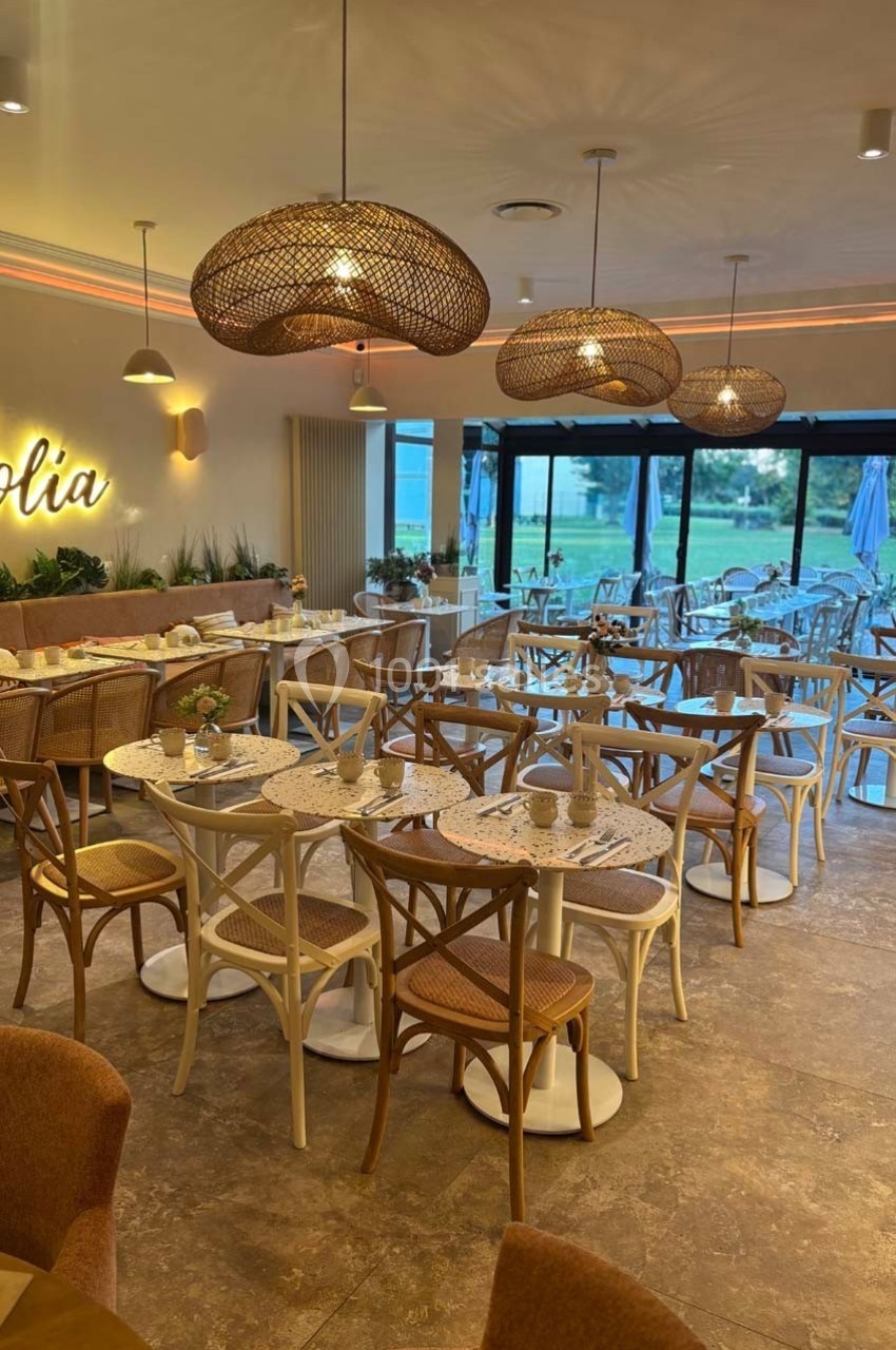 Salle de restaurant lumineuse avec tables dressées, chaises en bois et luminaires suspendus en osier.