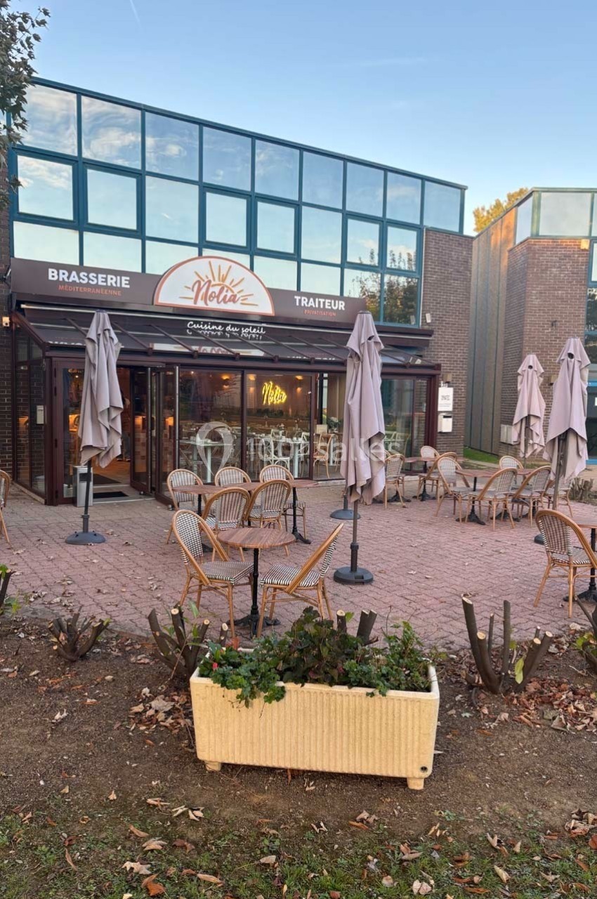 Terrasse d'une brasserie avec chaises en rotin, parasols fermés, devant une façade vitrée et un bâtiment moderne.