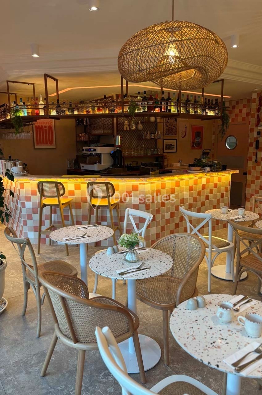 Intérieur d'un café avec des tables en terrazzo, des chaises en rotin et un bar éclairé entouré de carreaux colorés.