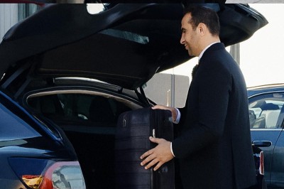 Un homme en costume charge une valise dans le coffre ouvert d'une voiture devant un bâtiment.