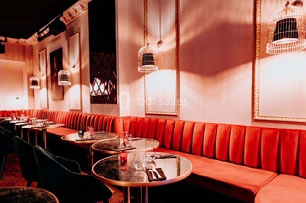 Salle de restaurant avec banquettes rouges, tables rondes en marbre noir et cages décoratives suspendues.