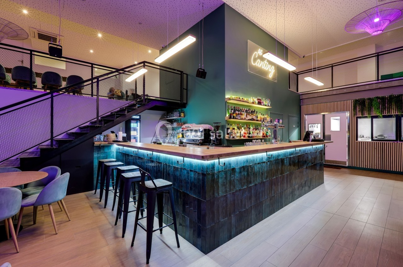 Bar moderne avec comptoir éclairé, tabourets hauts, étagères de bouteilles, escalier métallique et ambiance lumineuse.