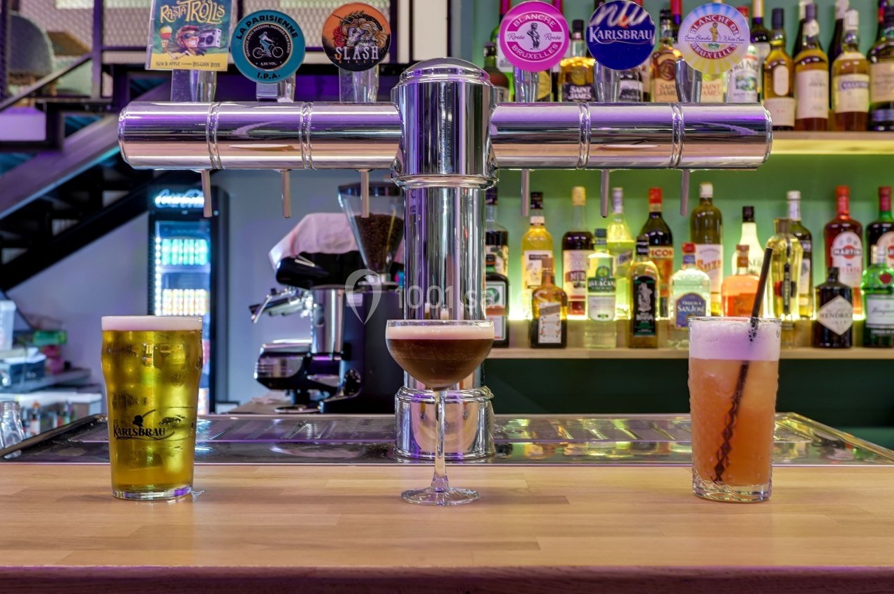 Trois verres de boissons différentes posés sur un comptoir de bar, avec des bouteilles et des tireuses en arrière-plan.