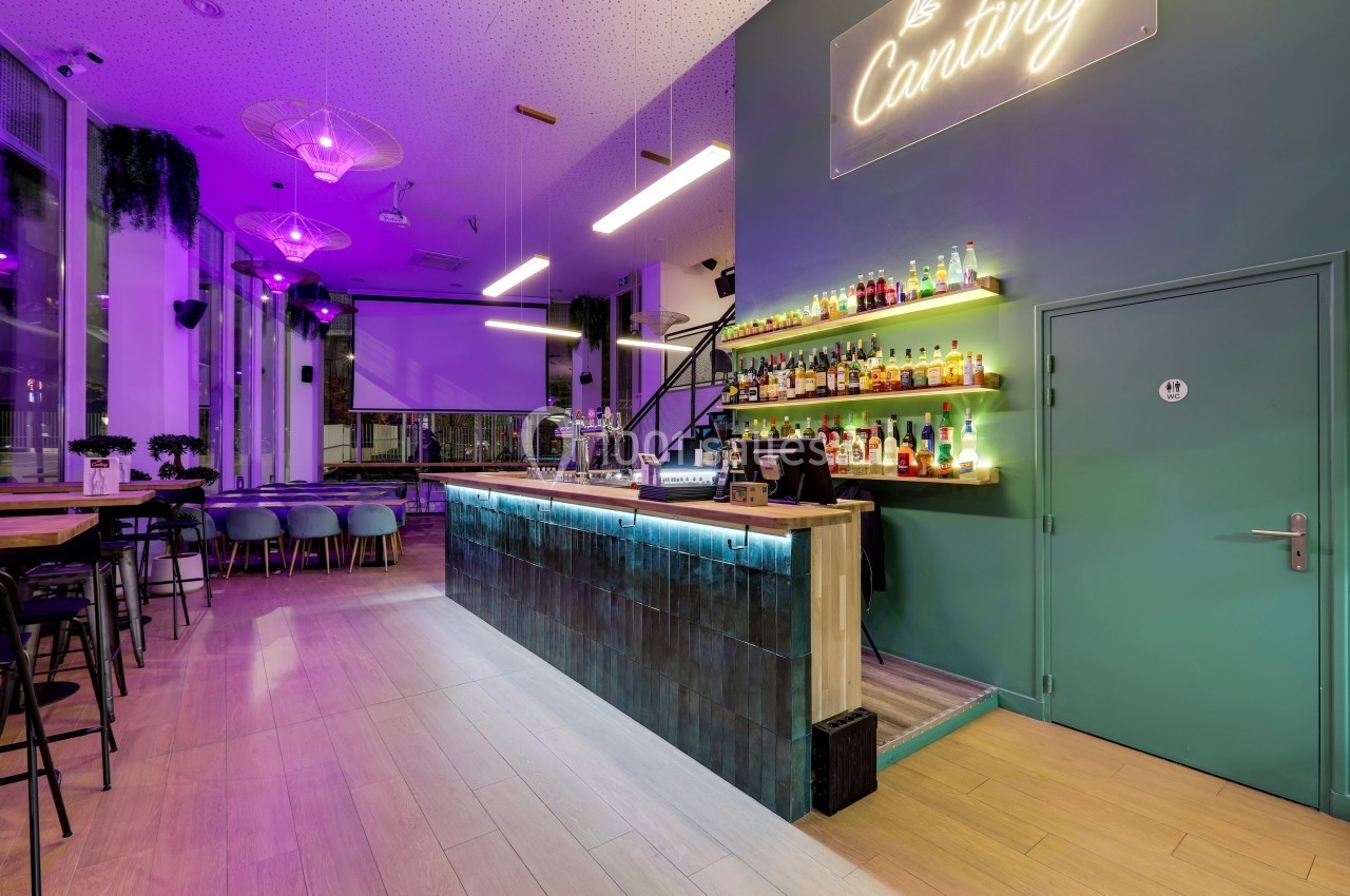 Bar moderne avec comptoir éclairé, étagères de bouteilles, tables et chaises, ambiance lumineuse violette.