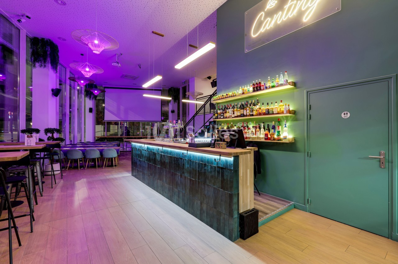 Bar moderne avec comptoir éclairé, étagères de bouteilles, tables et chaises, ambiance lumineuse violette.