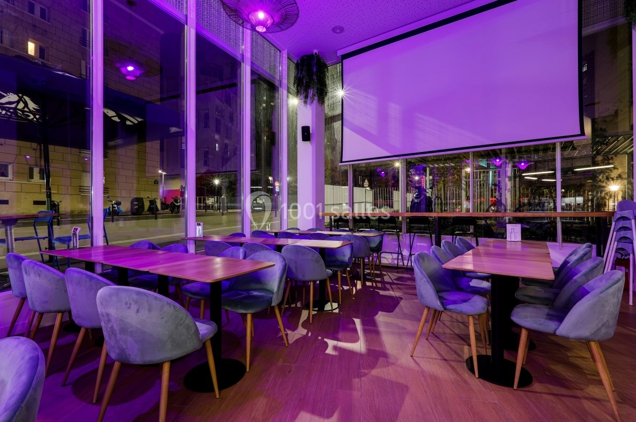 Salle de restaurant moderne avec tables en bois, chaises rembourrées et éclairage violet, donnant sur de grandes baies…