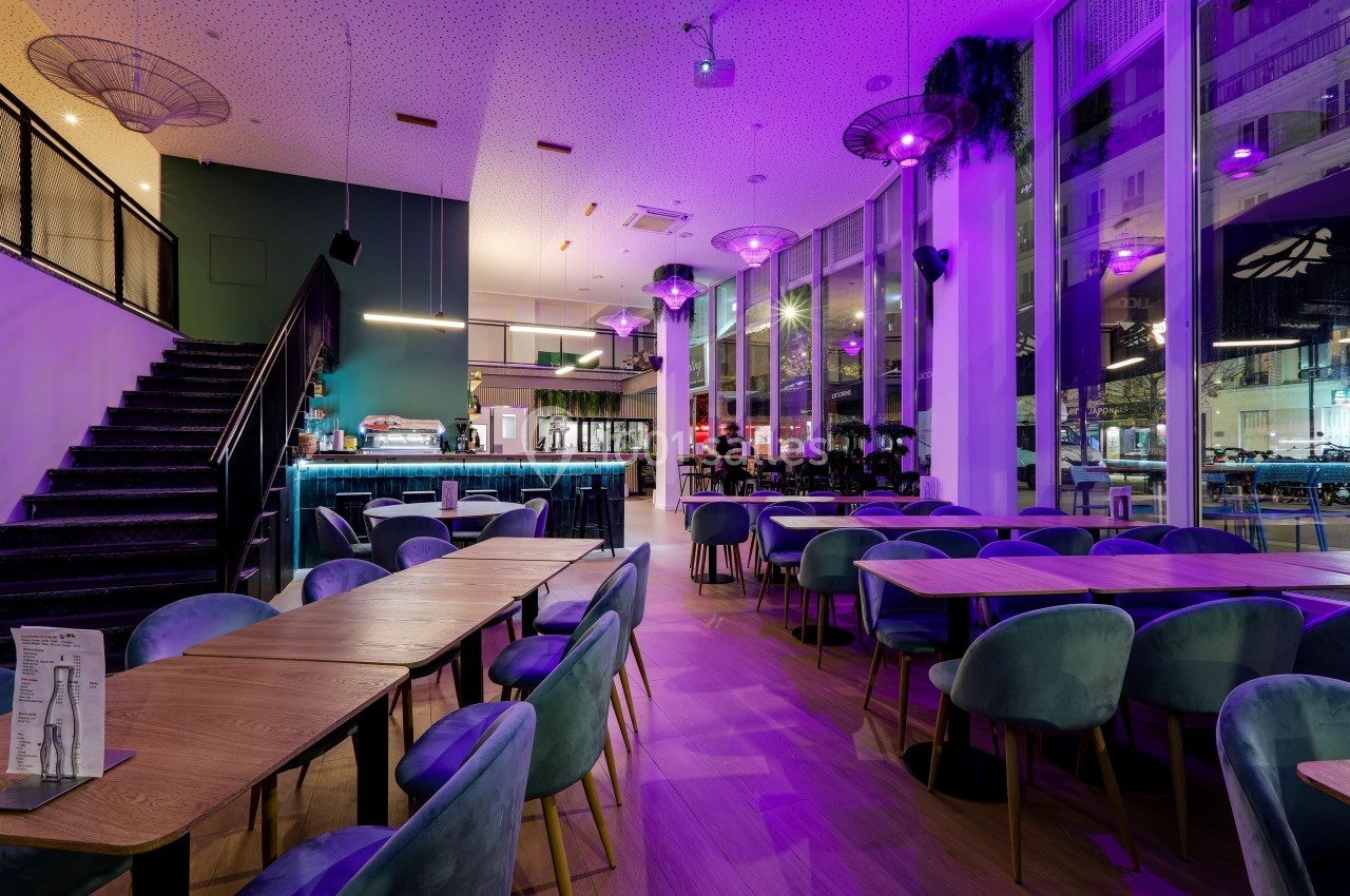 Salle de restaurant moderne avec grandes baies vitrées, éclairage violet et mobilier en bois et velours.