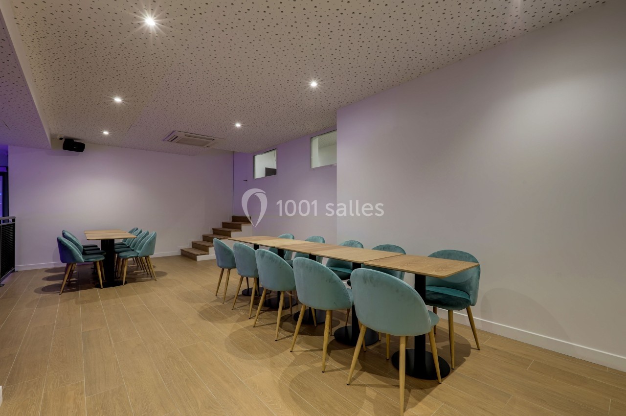 Salle moderne avec tables en bois, chaises bleu-vert, sol en parquet clair et plafond perforé éclairé par des spots.