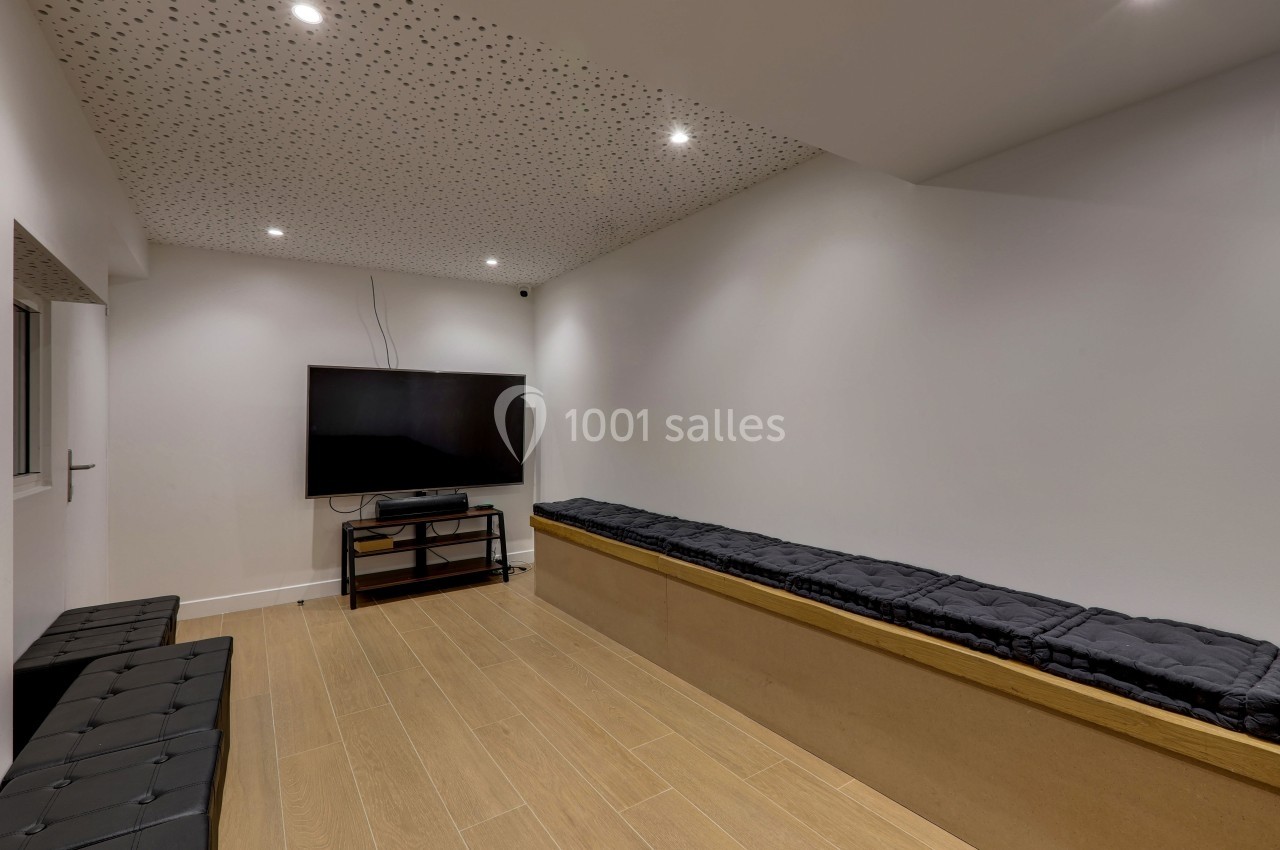Salle moderne avec un écran plat fixé sur un meuble, bancs rembourrés et plafond perforé éclairé par des spots.
