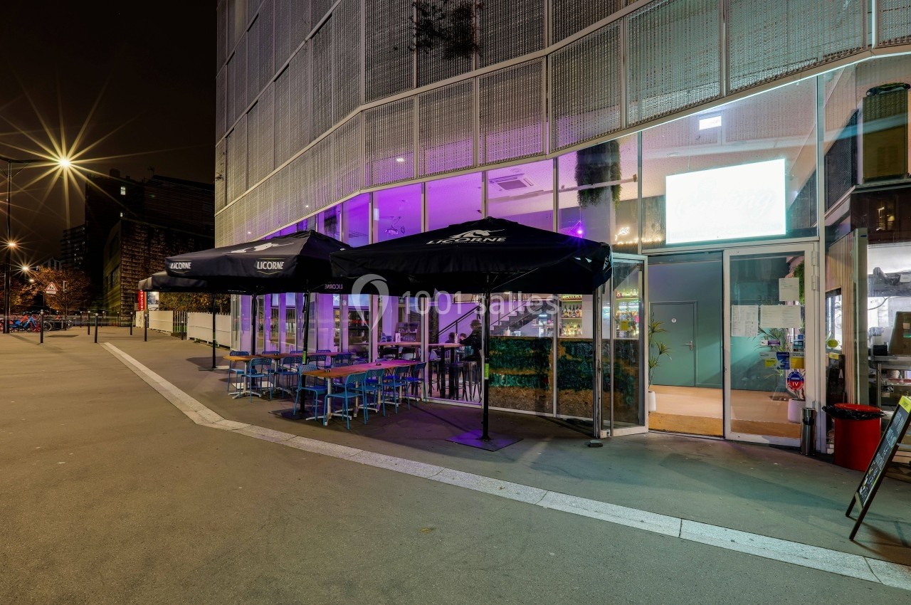 Terrasse extérieure d'un café-restaurant éclairée la nuit, avec tables, chaises et parasols noirs.
