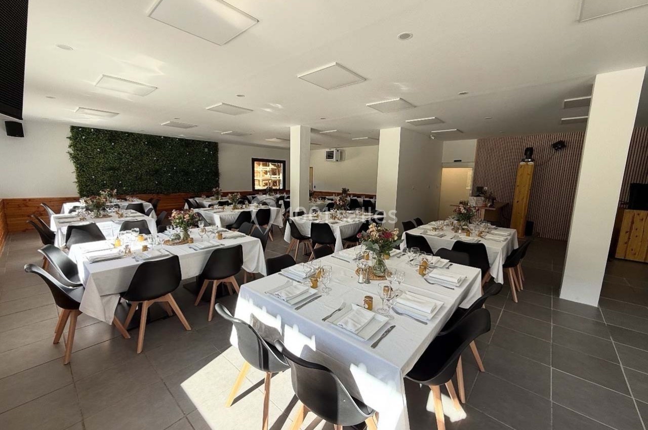 Salle lumineuse avec tables dressées pour un événement, décorée de fleurs et chaises noires modernes.