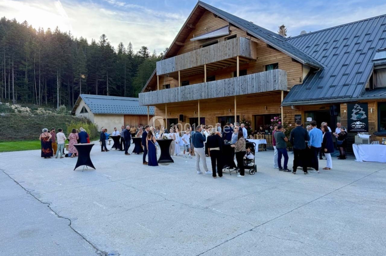 Groupe de personnes rassemblées devant un grand chalet en bois, dans un cadre naturel avec forêt en arrière-plan.