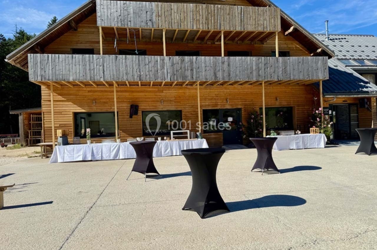 Chalet en bois avec terrasse, tables hautes noires et nappes blanches disposées sur une grande esplanade ensoleillée.