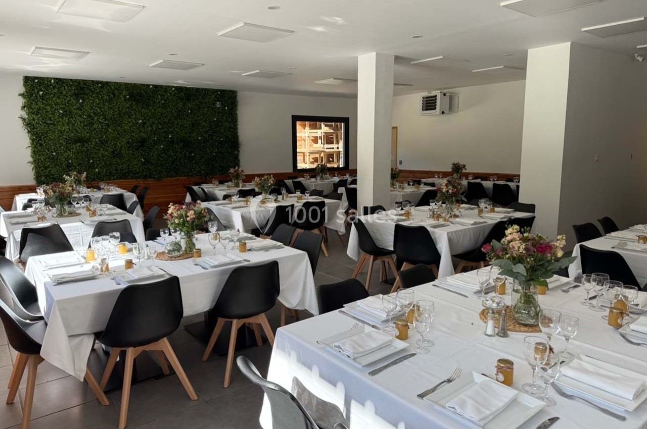 Salle de réception lumineuse avec tables dressées, nappes blanches, chaises noires et décoration florale.