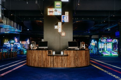 Comptoir en bois avec deux ordinateurs dans une salle d'arcade lumineuse, entourée de jeux et de machines.