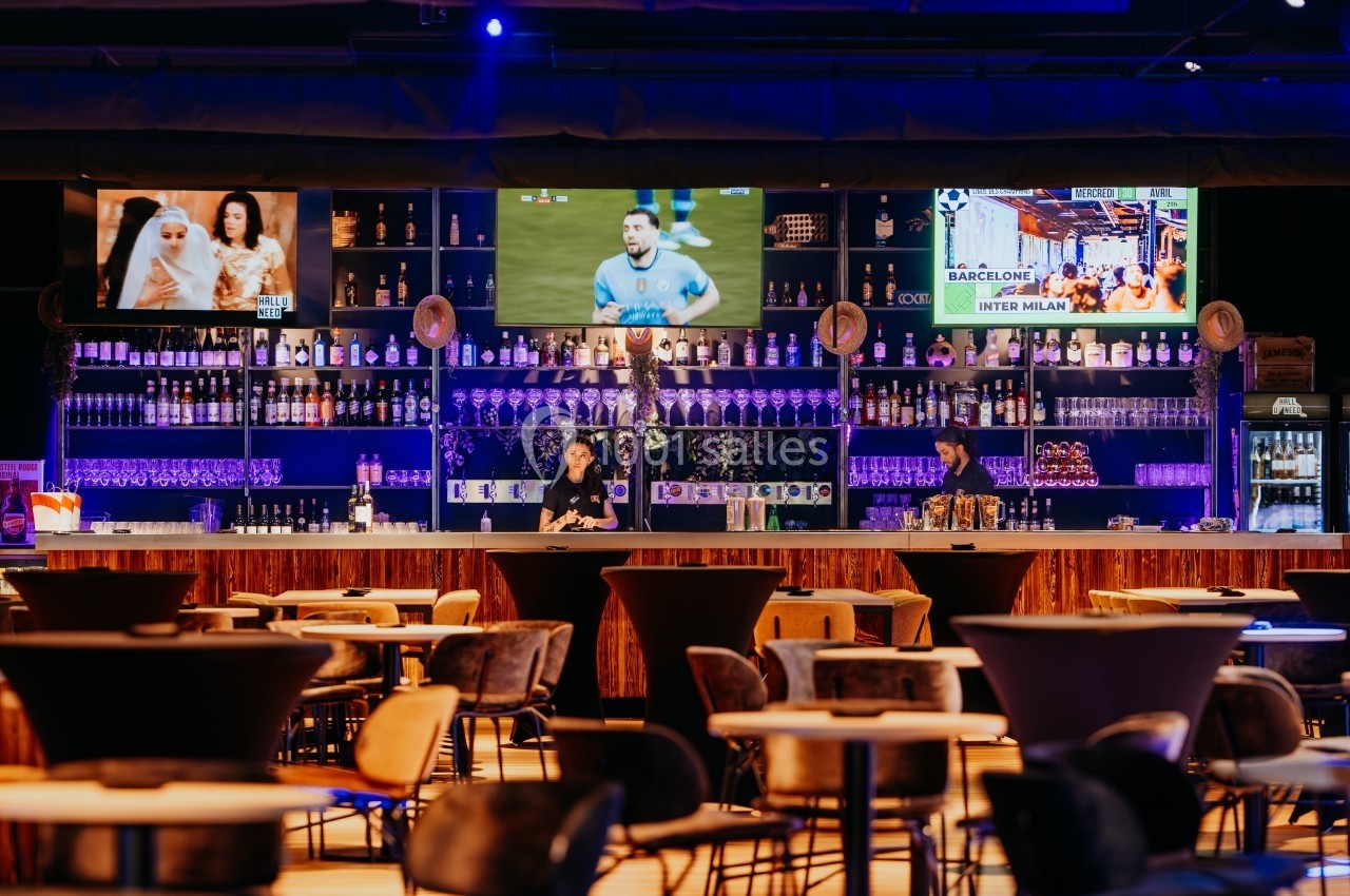 Bar moderne avec étagères remplies de bouteilles, tables vides au premier plan et écrans diffusant des vidéos en arrière…