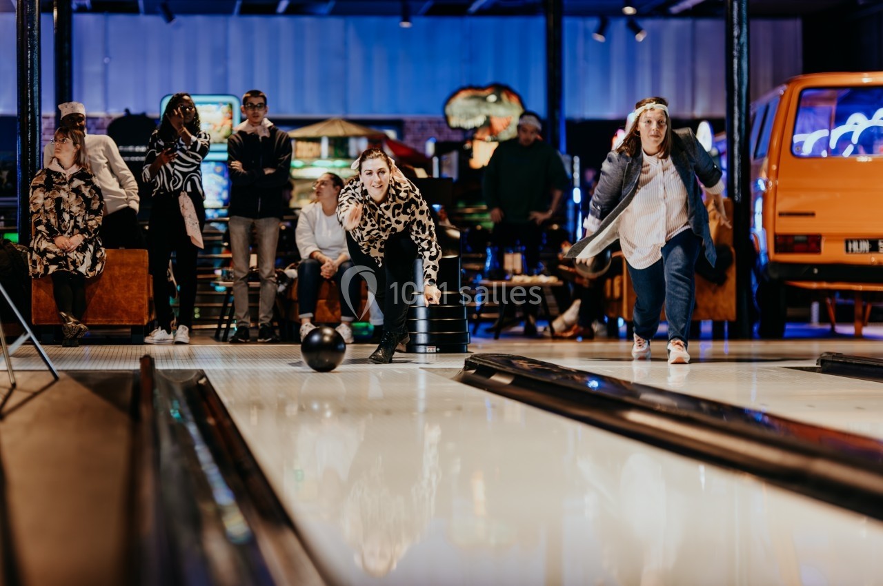 Deux femmes jouent au bowling dans une salle animée, avec des spectateurs et des lumières colorées en arrière-plan.