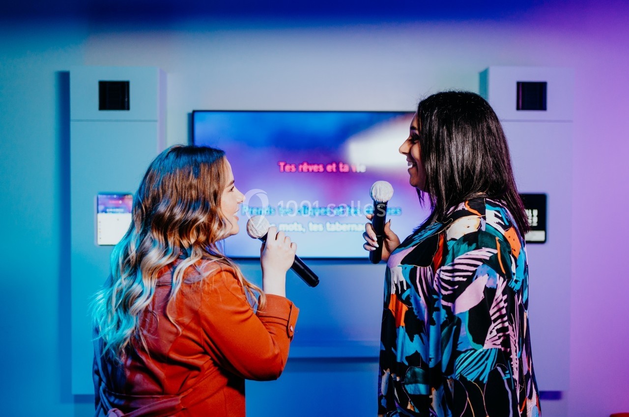 Deux femmes chantent au karaoké devant un écran affichant les paroles d'une chanson, dans une ambiance colorée.