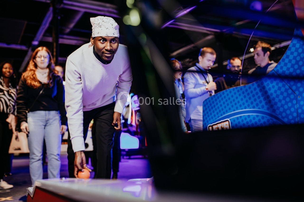 Un homme joue à un jeu d'adresse dans une salle d'arcade, observé par un groupe de personnes en arrière-plan.