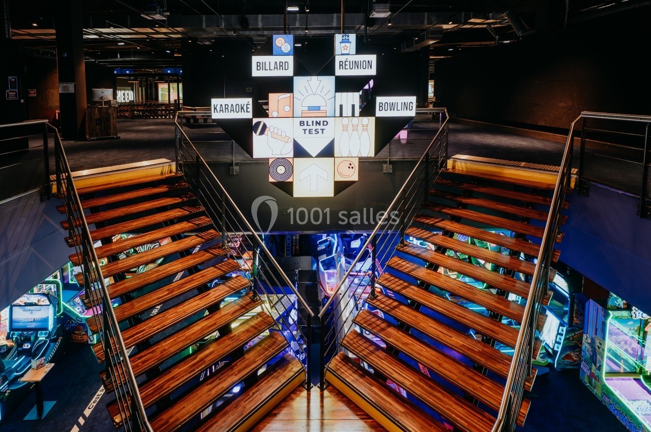 Escalier en bois menant à un espace de loisirs avec panneaux lumineux indiquant diverses activités comme billard et karaoké.