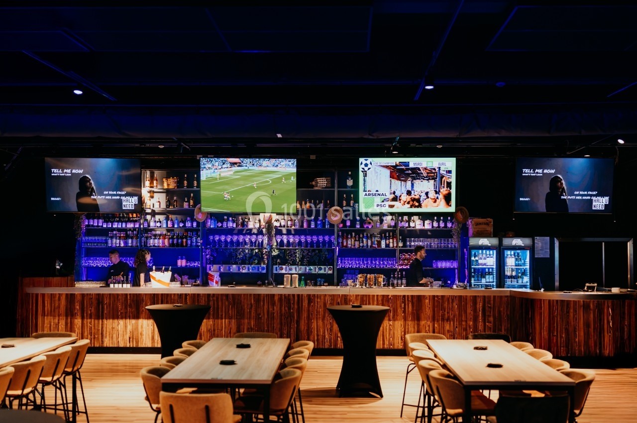 Bar moderne avec comptoir éclairé, tables en bois, écrans diffusant des vidéos et étagères remplies de bouteilles.