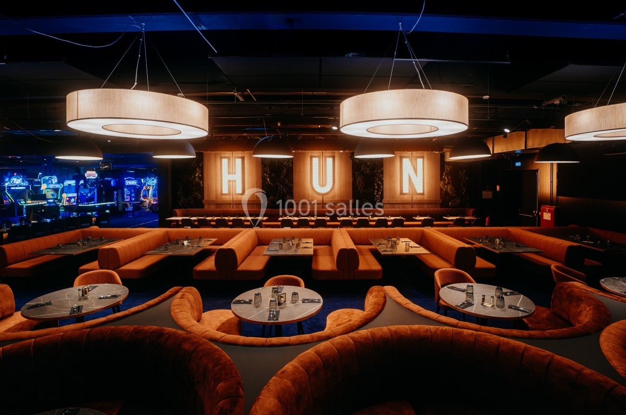 Salle de restaurant avec banquettes orange, tables rondes et grandes lettres lumineuses formant ’HUN’ en arrière-plan.