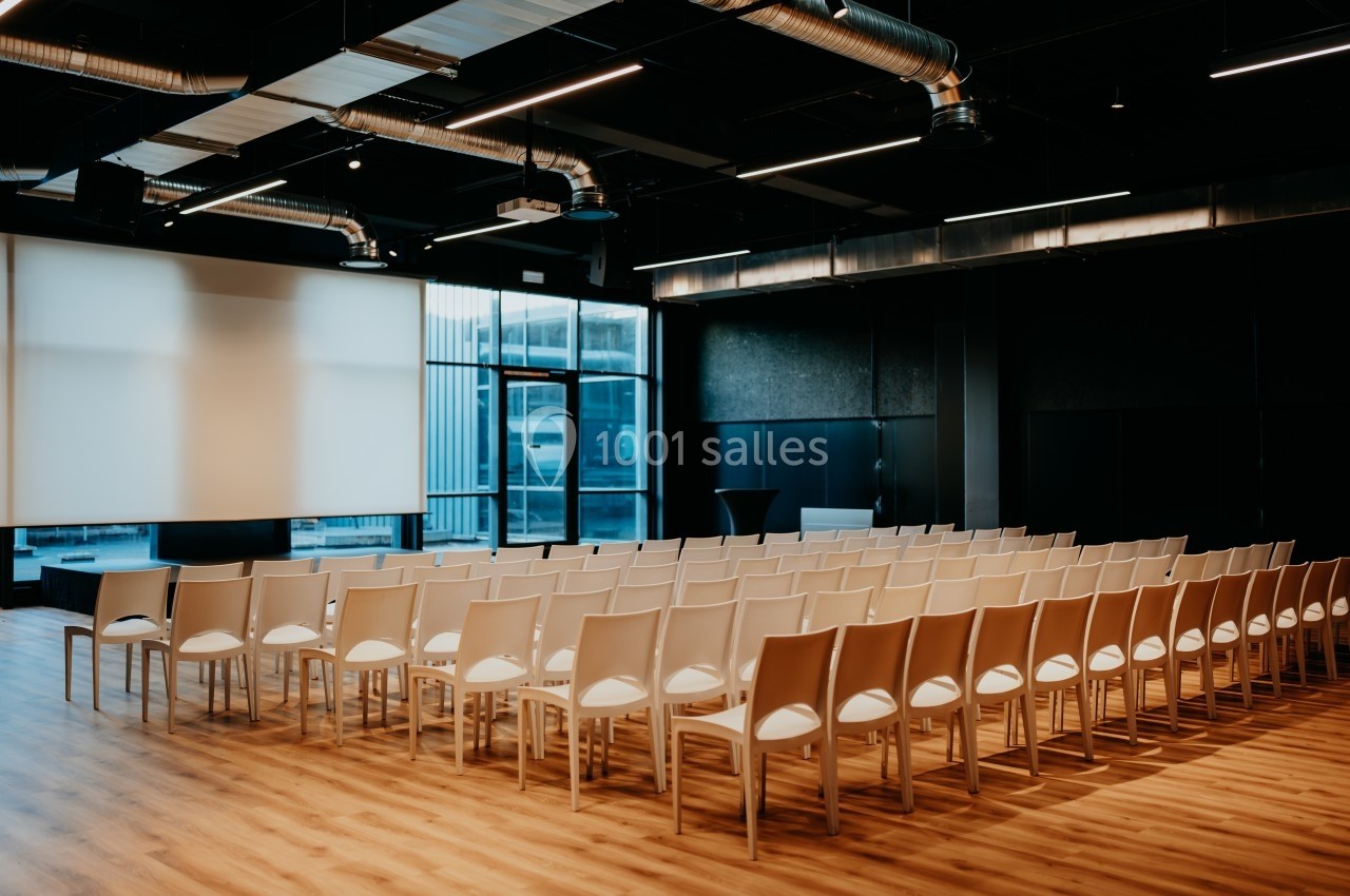 Salle de conférence moderne avec des rangées de chaises blanches alignées face à un écran de projection.