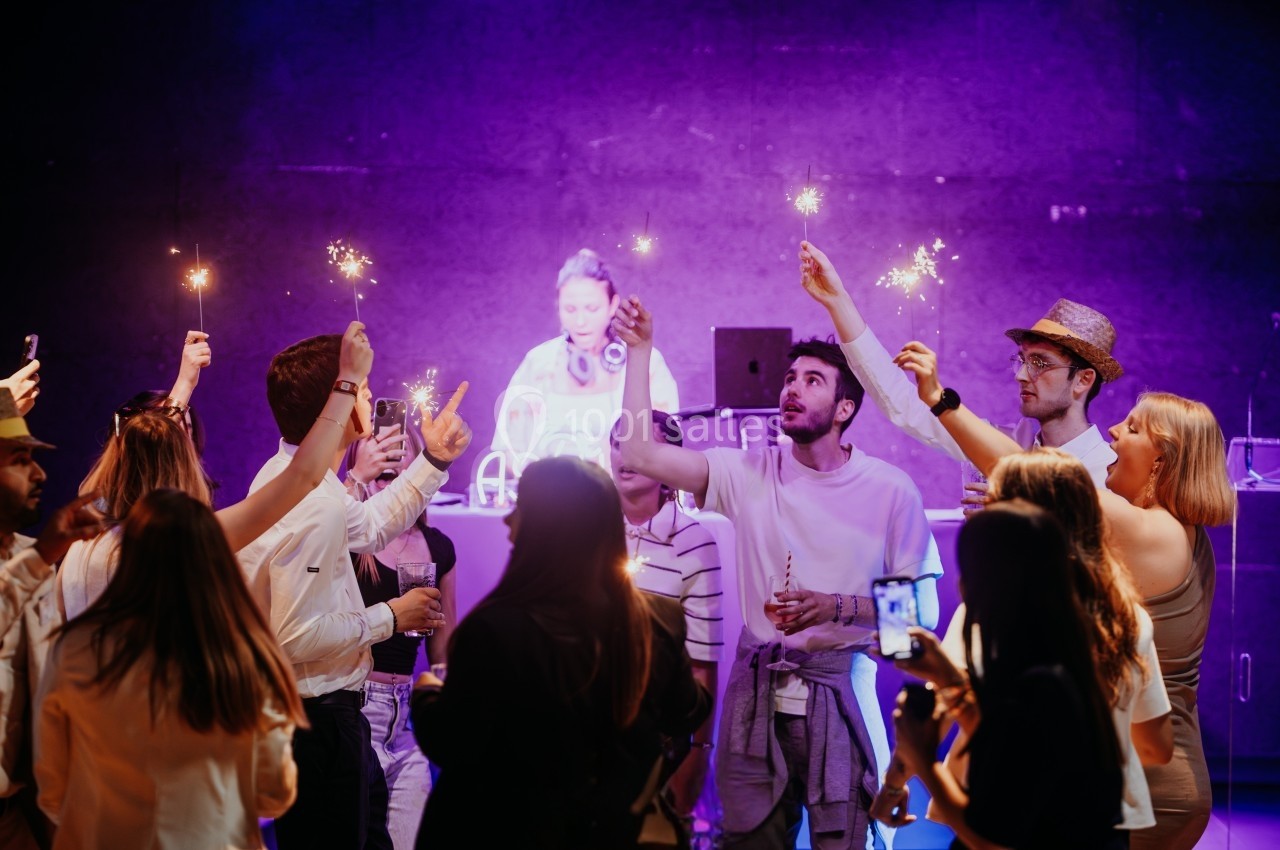 Groupe de personnes célébrant avec des cierges magiques devant un DJ sur une scène éclairée par des lumières violettes.