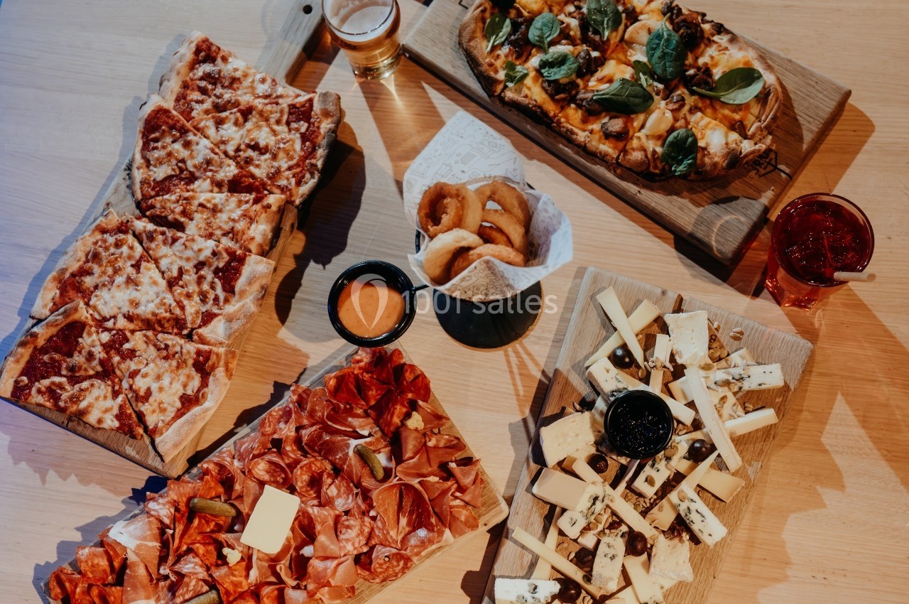 Plateaux de charcuterie, fromages, pizzas et beignets servis avec des sauces et des boissons sur une table en bois.