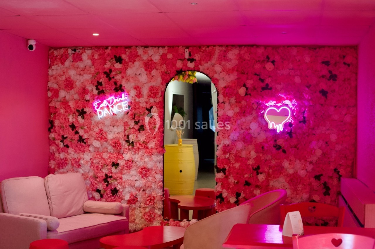 Mur couvert de fleurs roses avec des néons en forme de cœur et texte, mobilier rose et fauteuil beige au premier plan.