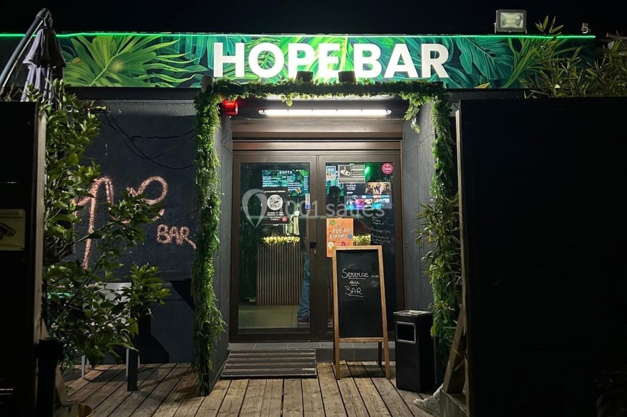 Façade d'un bar avec enseigne lumineuse ’Hope Bar’, décorée de plantes et d'une ardoise à l'entrée.