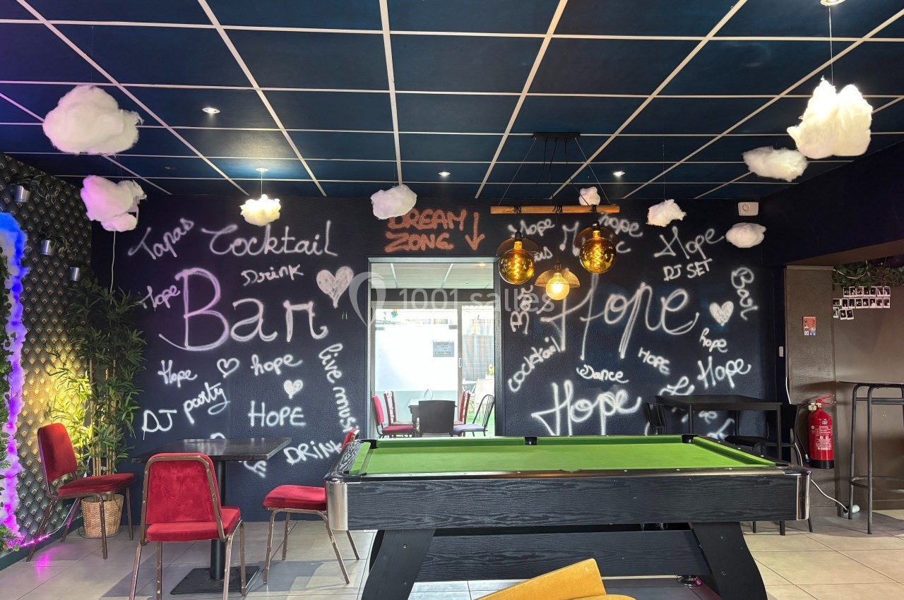 Salle de loisirs avec billard, murs décorés de graffitis et nuages suspendus sous un plafond bleu.