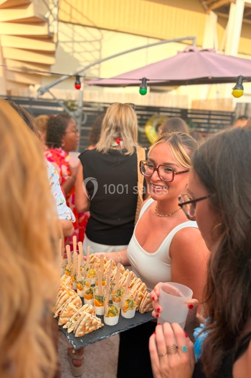 Une femme souriante tient un plateau de verrines et amuse-bouches lors d'un événement en extérieur.