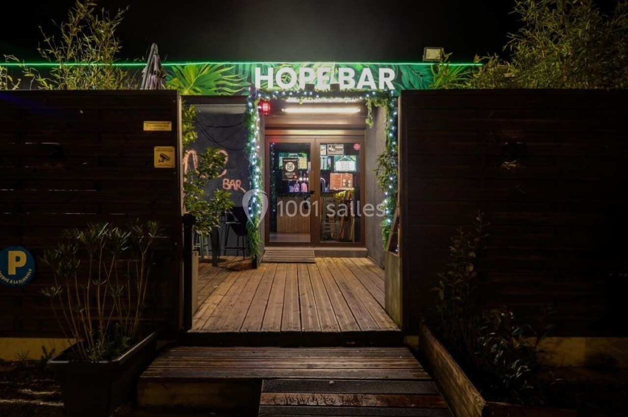 Entrée d'un bar en bois éclairé la nuit, avec une enseigne lumineuse ’Hop Bar’ et une terrasse entourée de végétation.