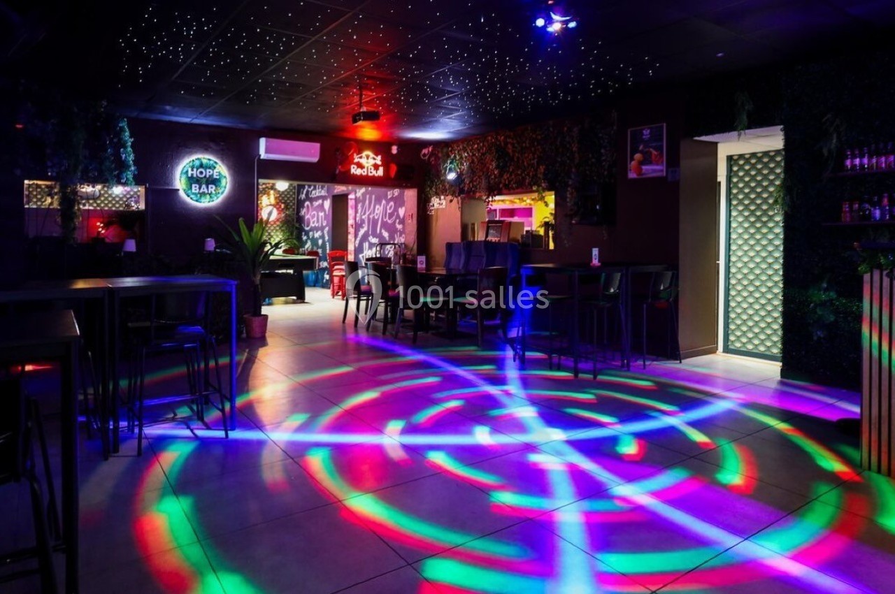 Salle de bar avec éclairage coloré au sol, décor végétal et enseignes lumineuses sur les murs.