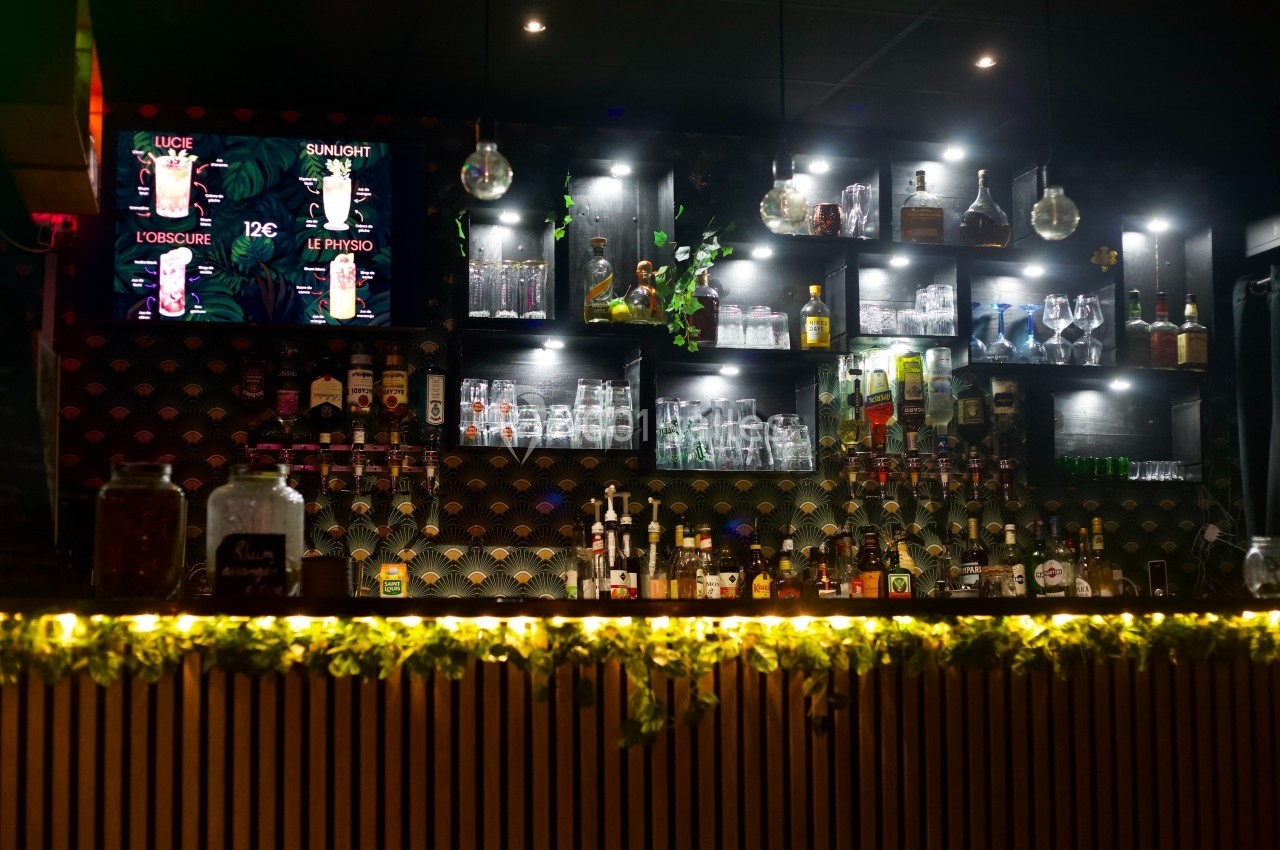 Bar avec étagères éclairées contenant des bouteilles et verres, écran affichant des cocktails, décoration végétale en façade.