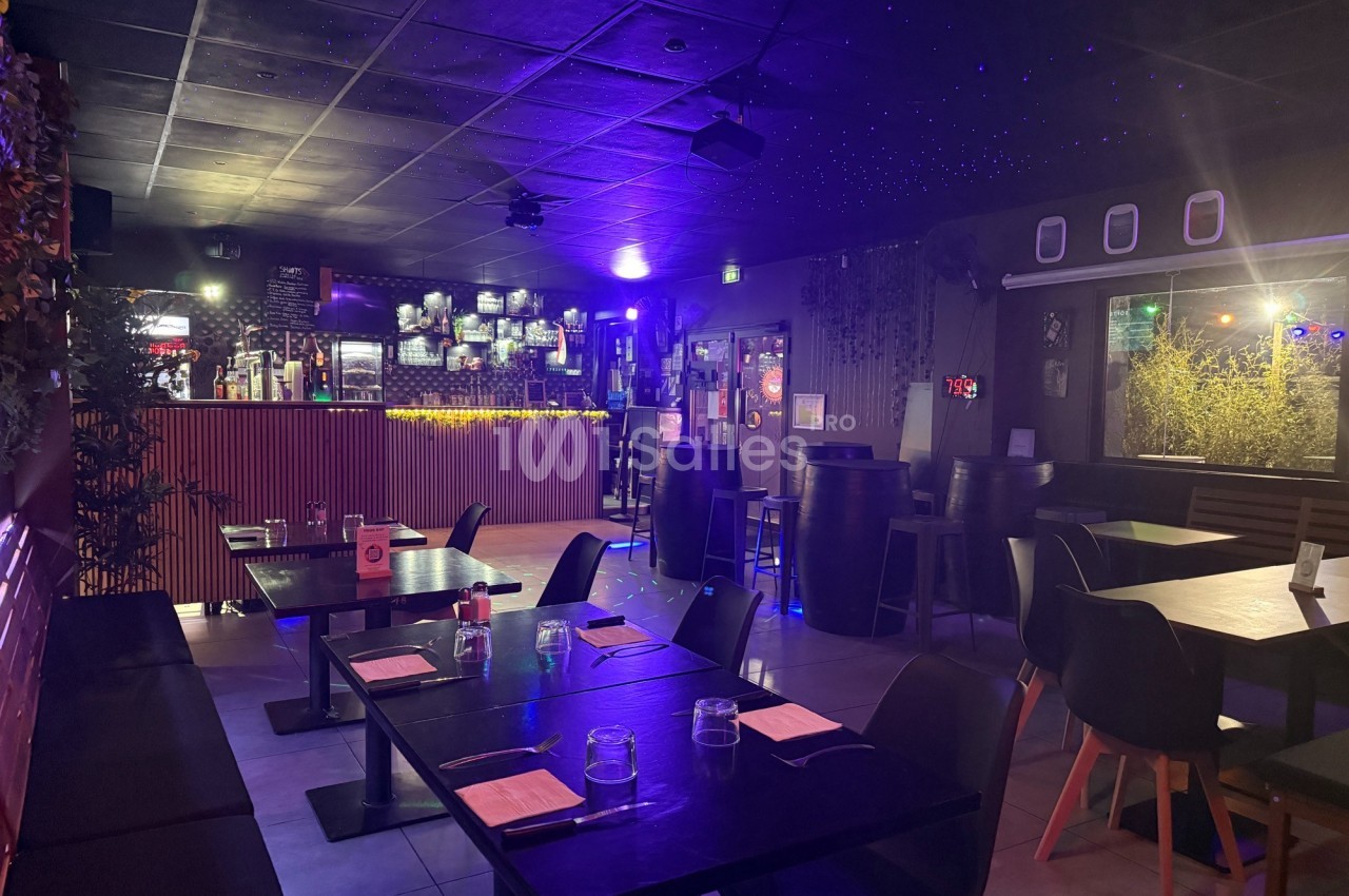 Salle de restaurant avec tables dressées, éclairage tamisé et bar illuminé en arrière-plan.