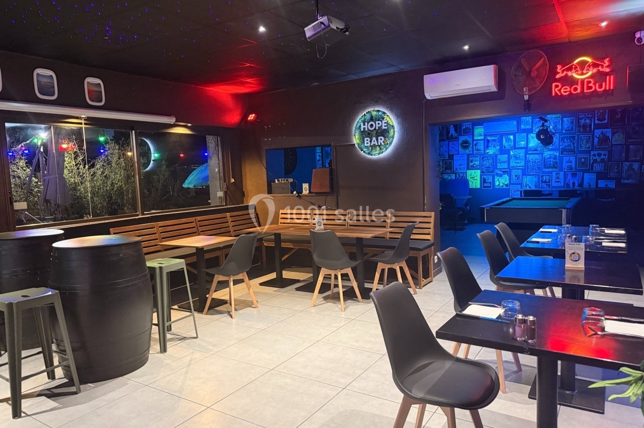 Salle de restaurant moderne avec tables, chaises, tonneaux en guise de tables hautes et éclairage tamisé coloré.